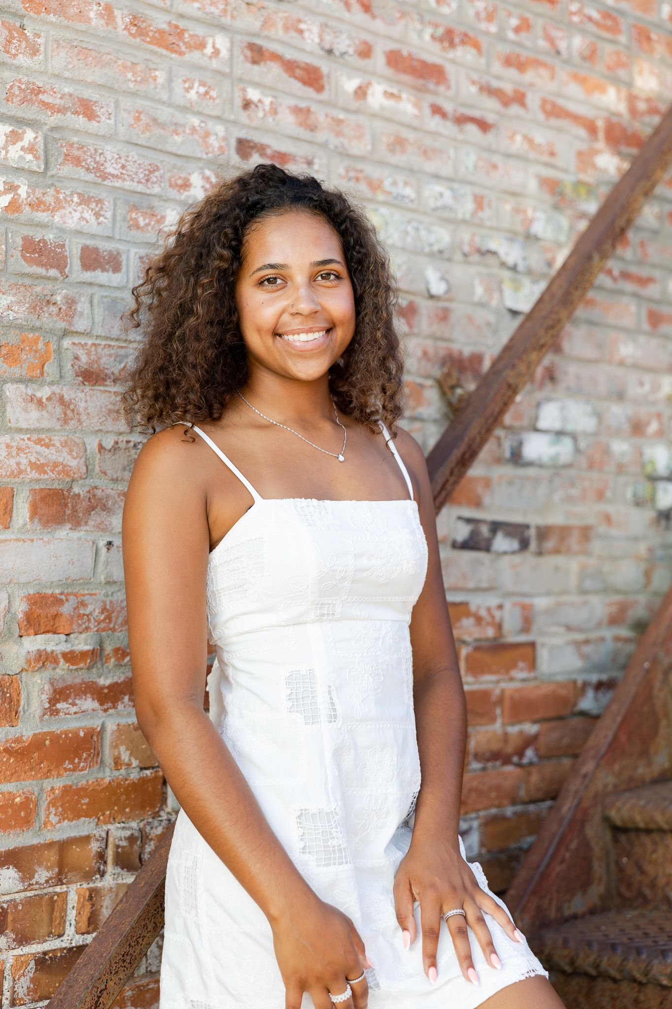 Person wearing a white sleeveless top poses against a rustic brick wall with a warm, welcoming smile.