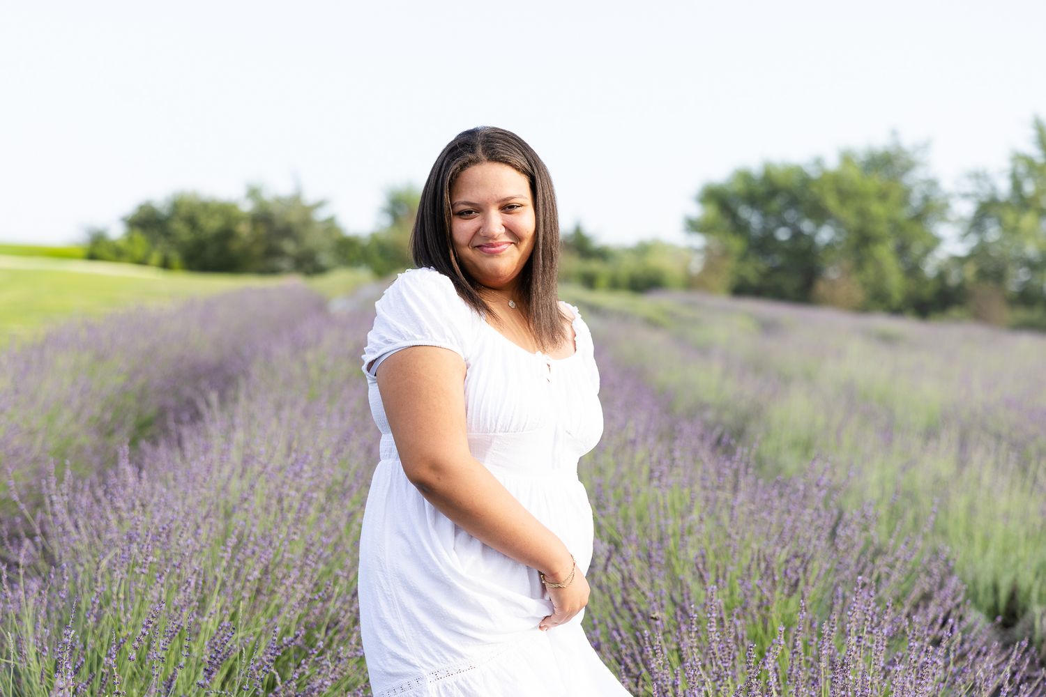 A person in a white dress poses in a scenic lavender field on a bright summer day.