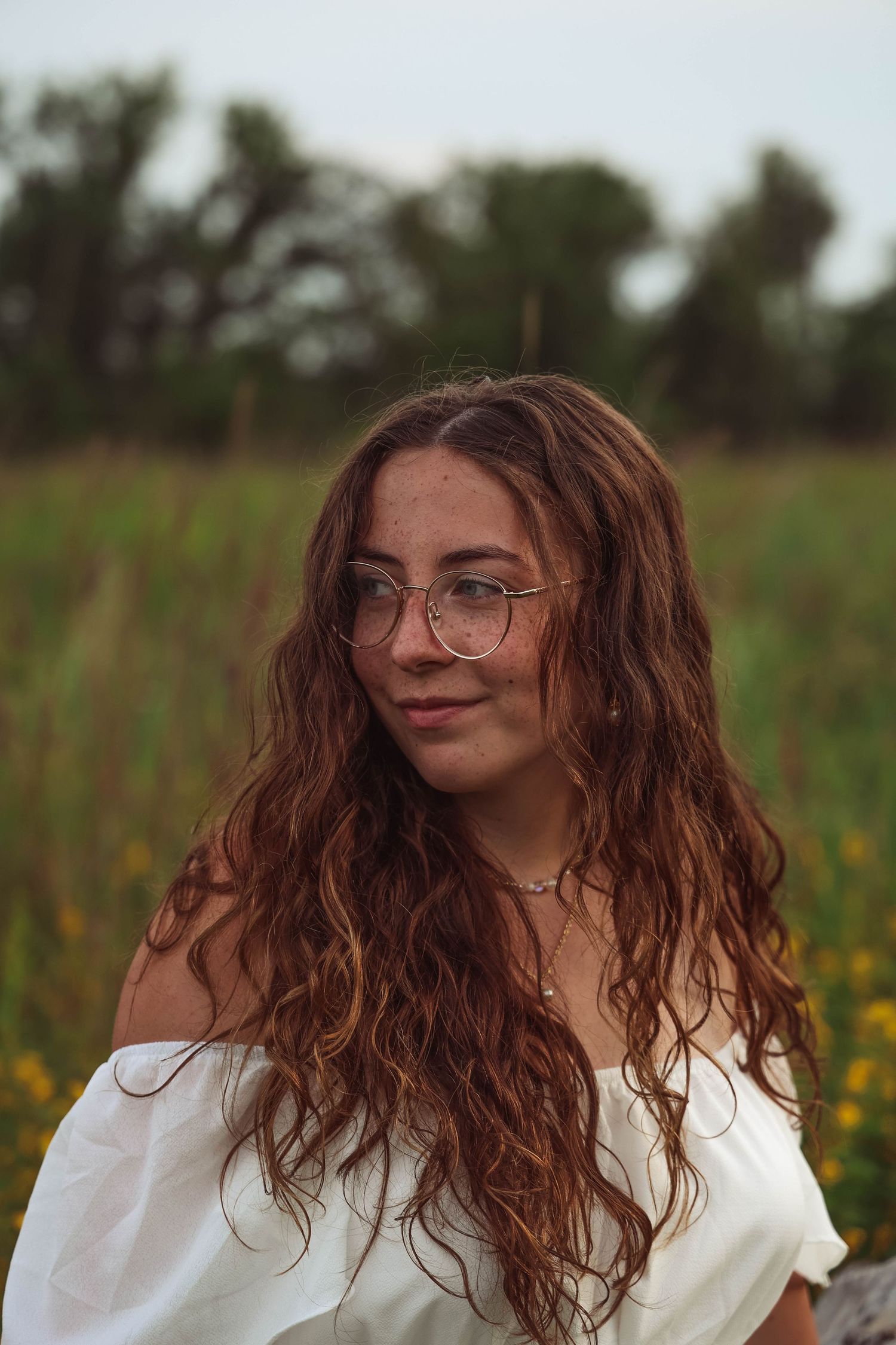 A portrait of someone with long wavy brown hair wearing glasses and a white off-shoulder top against a natural field background.