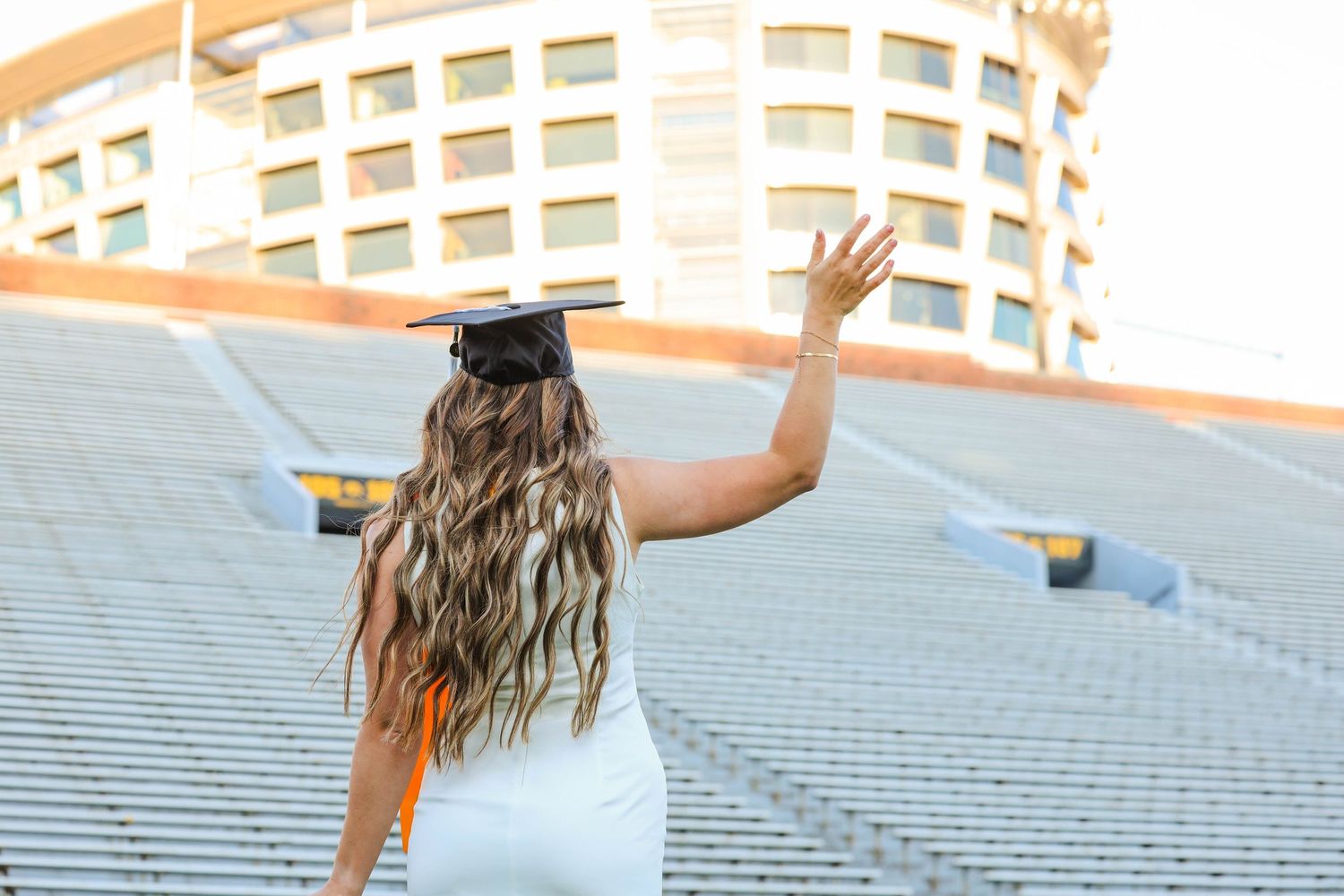 Graduate in white dress with long wavy hair waves farewell in Kinnick Stadium wearing cap and gown.