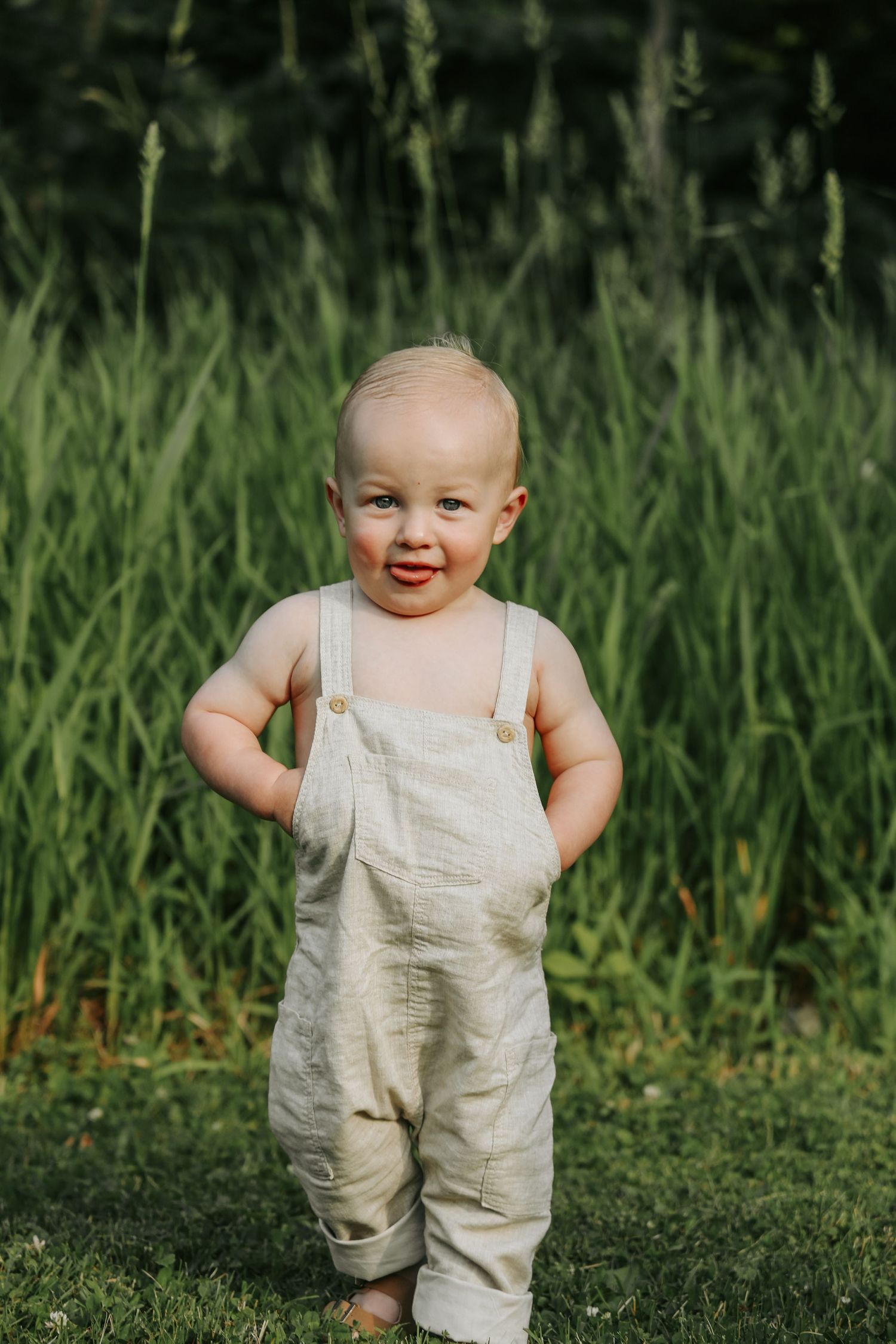 Toddler in linen overalls smiles while standing in tall grass.