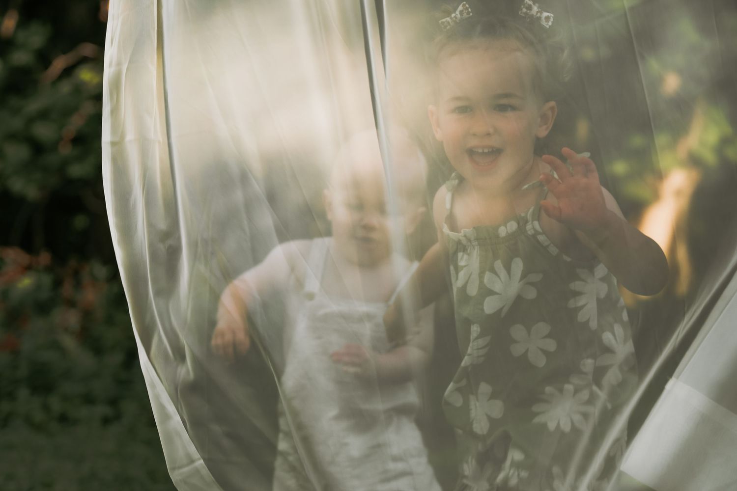 Children laugh behind sheer curtain creating dreamy outdoor portrait.