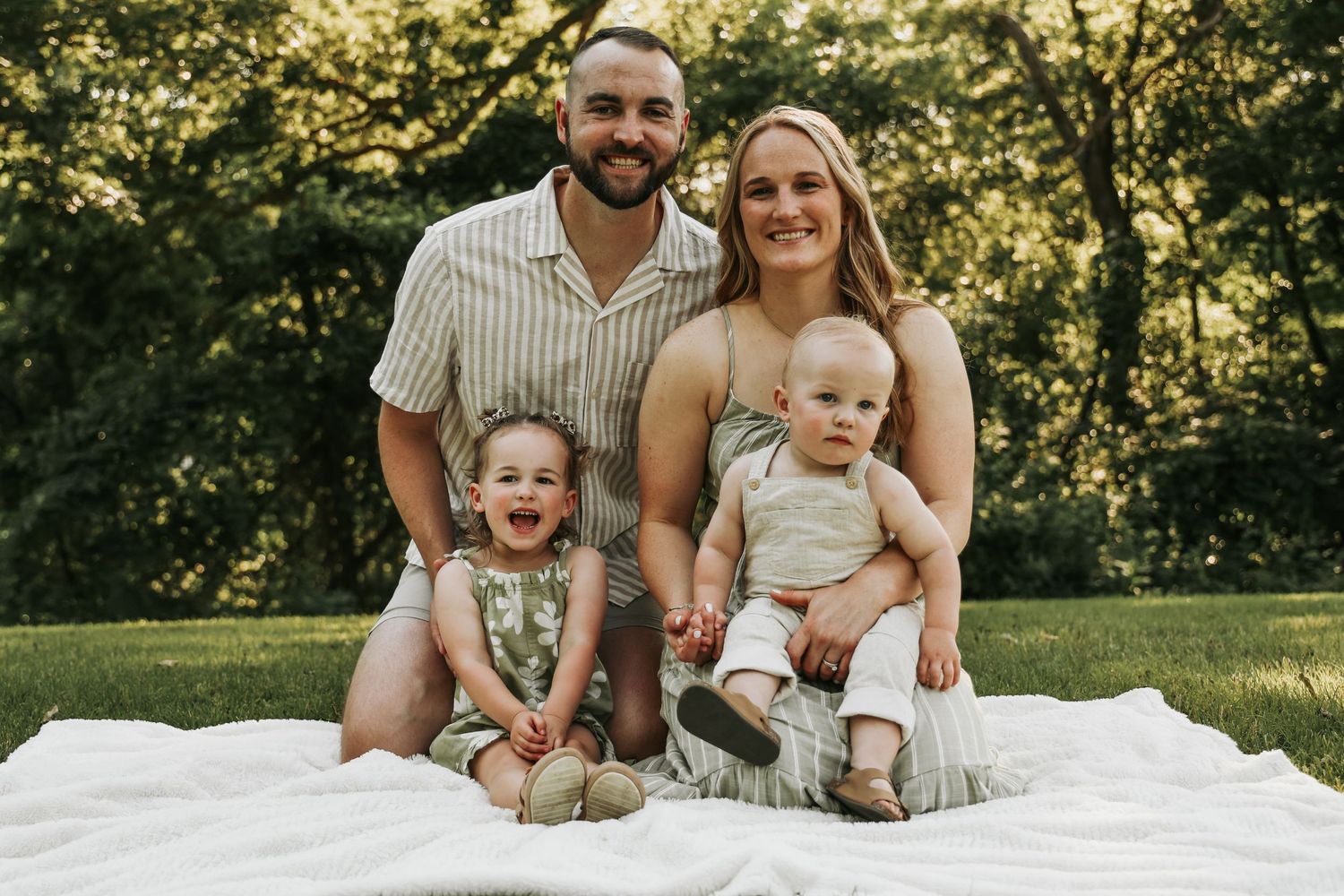 Happy family of four sits together on white blanket in sunny park setting.