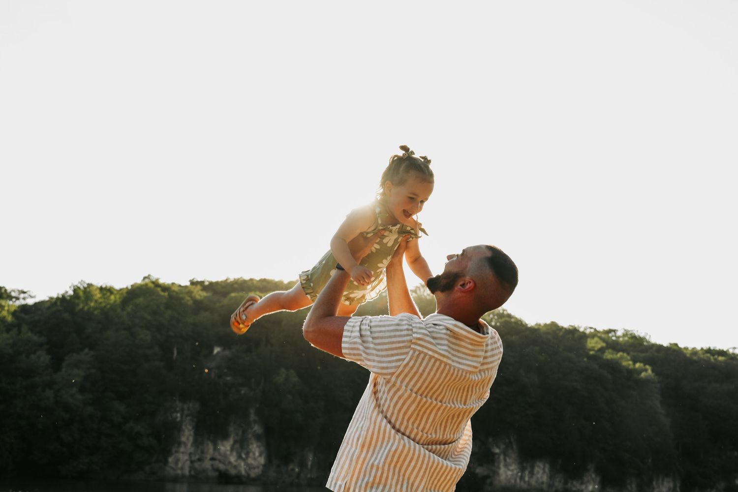 Parent lifts child playfully against sunset backdrop near cliffside.