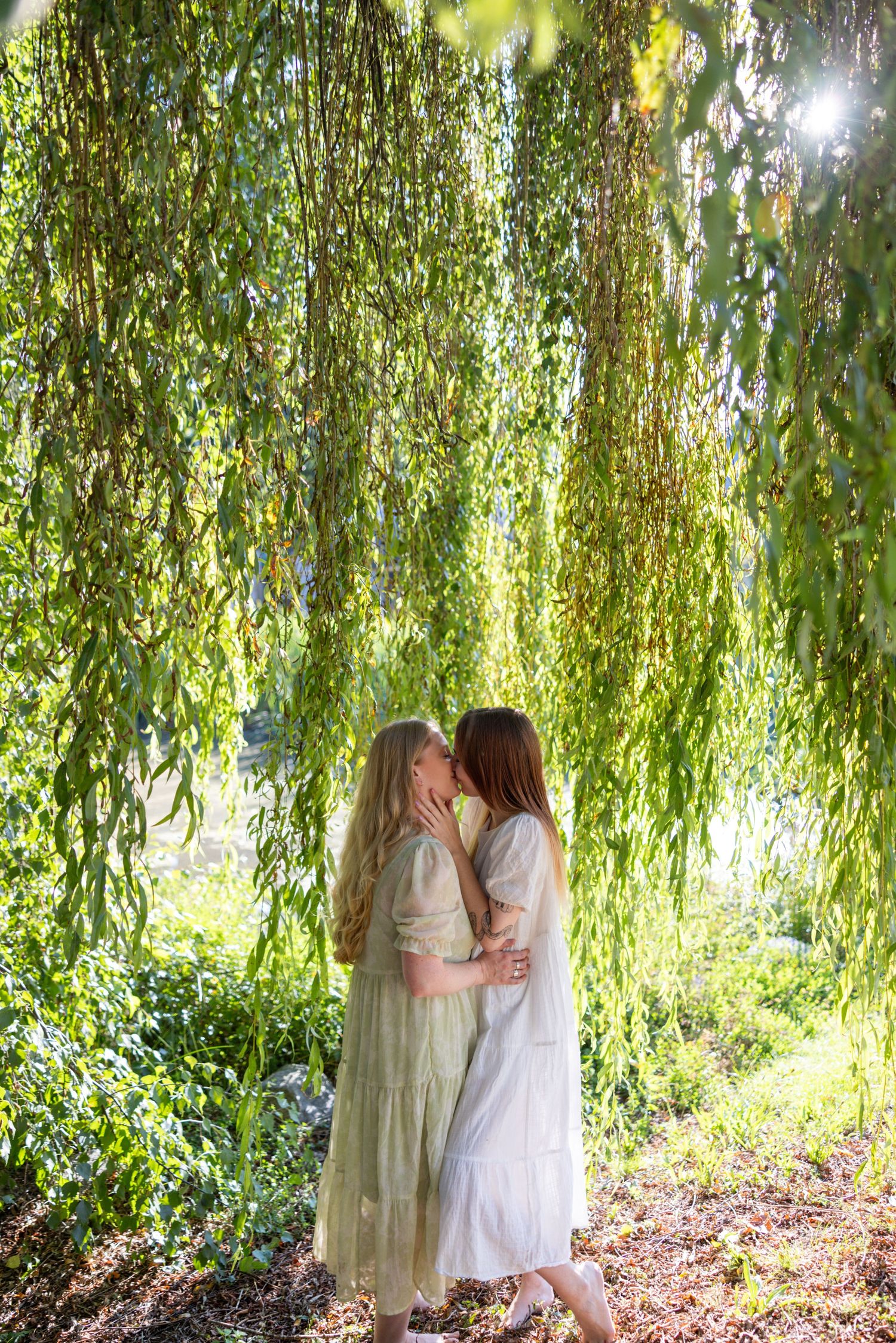 A couple embraces under drooping willow branches in dappled sunlight.