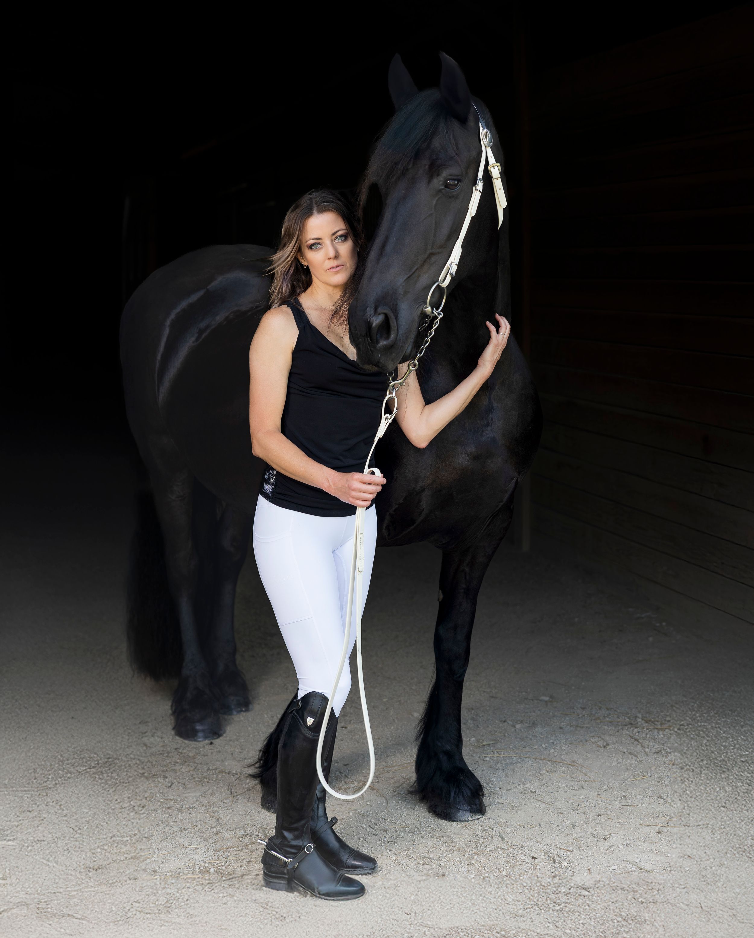 A woman in white pants stands next to a Friesian horse in a dark setting. Buhler Kansas, Black Gold Farm