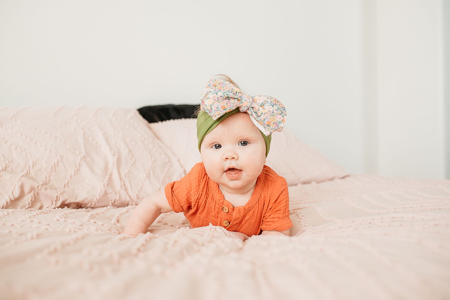Baby wearing orange onesie and bow headband lying on pink bedding.