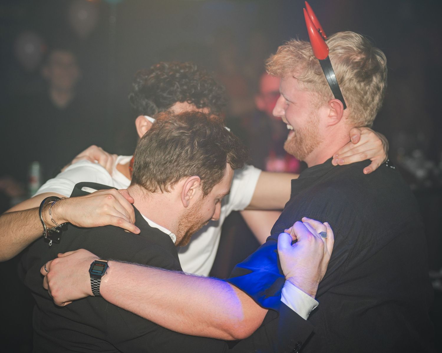 People energetically dancing and embracing at a nightclub with blue lighting effects in the background.