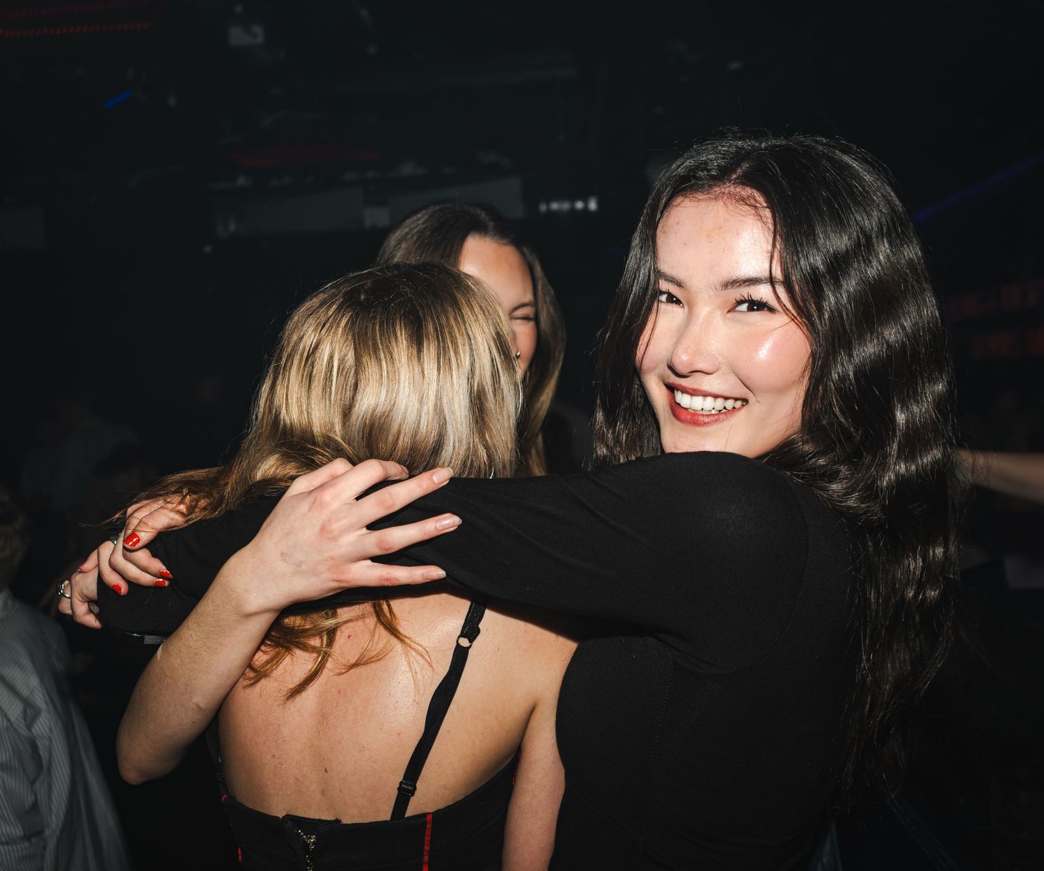 Friends embrace and laugh together at a dimly lit nightclub or party.