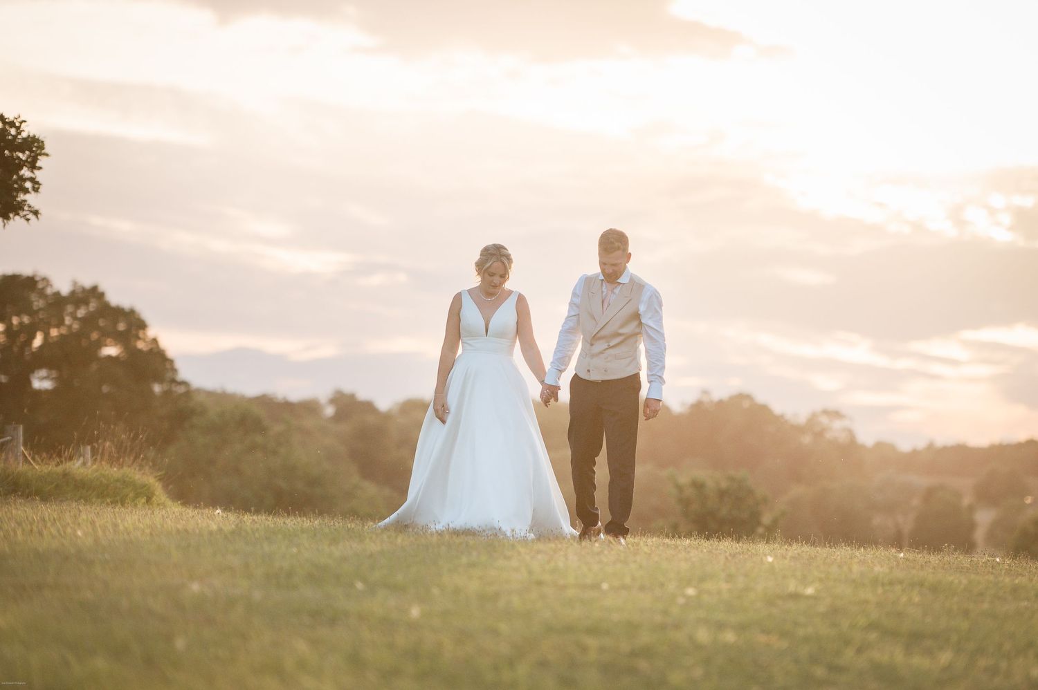 Newlyweds walking together at sunset across a hilltop meadow with mountains in the distance.
