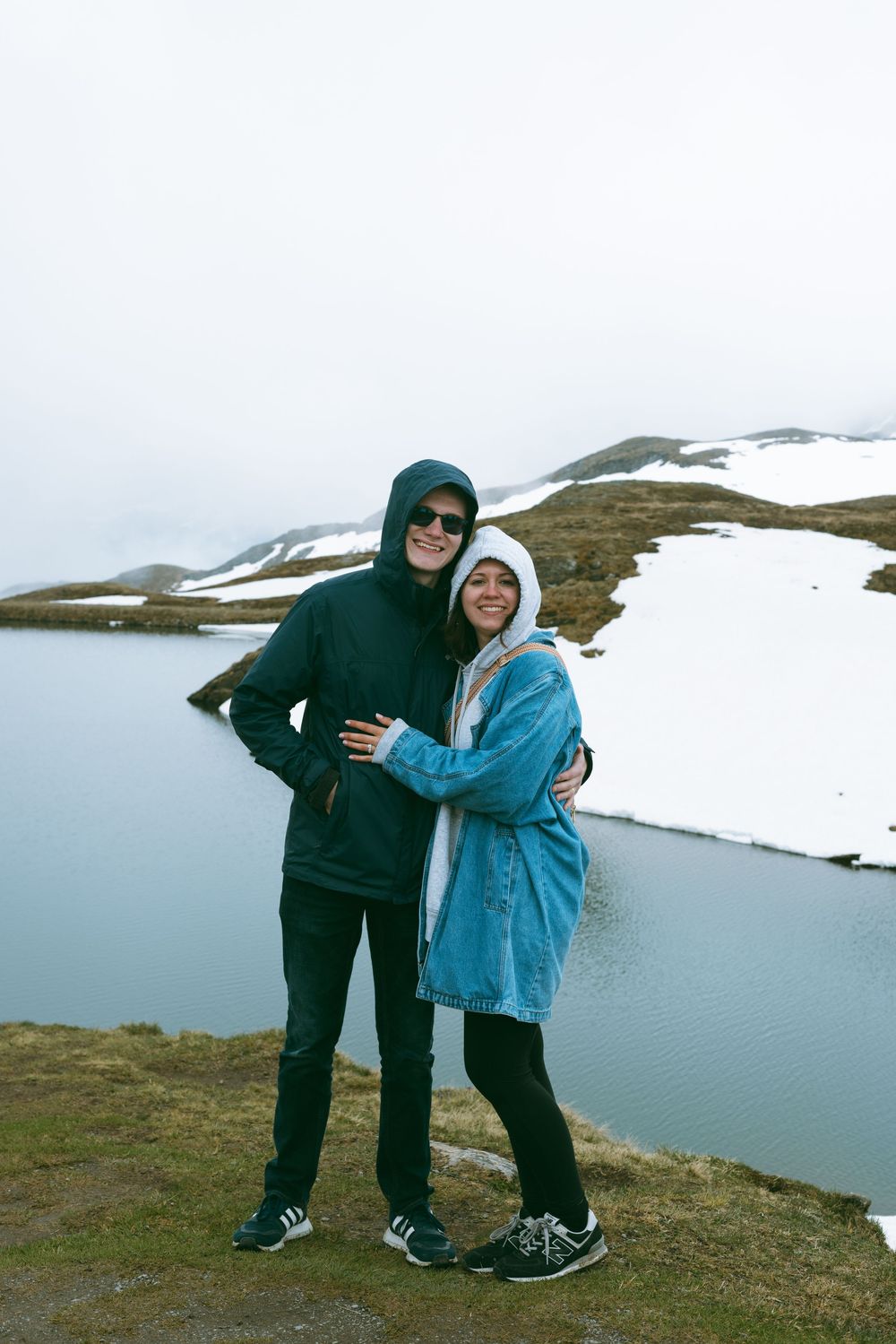 Two people in winter clothing embrace while standing on a snowy hillside with a lake view in the background.
