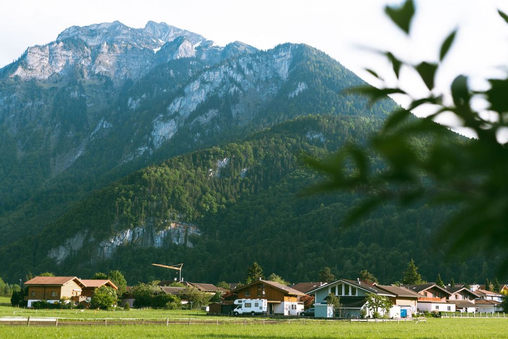 Alpine village nestled at the base of towering mountains with scattered houses and green meadows in the foreground.