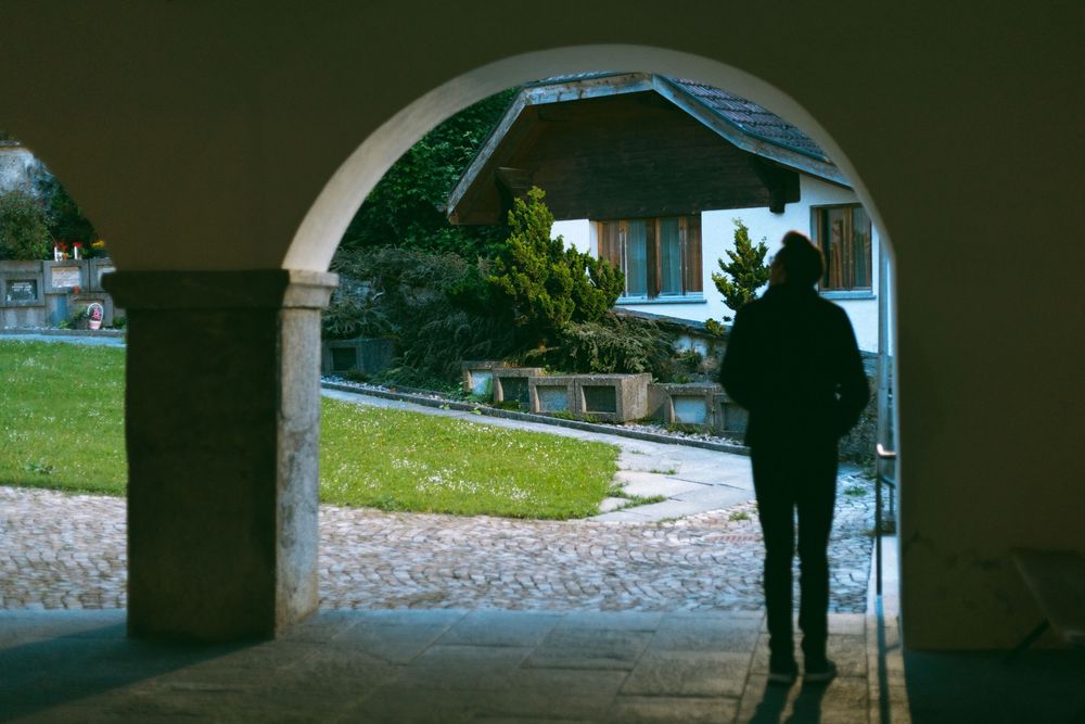 A silhouetted figure stands beneath a stone archway overlooking a grassy lawn and building in daylight.