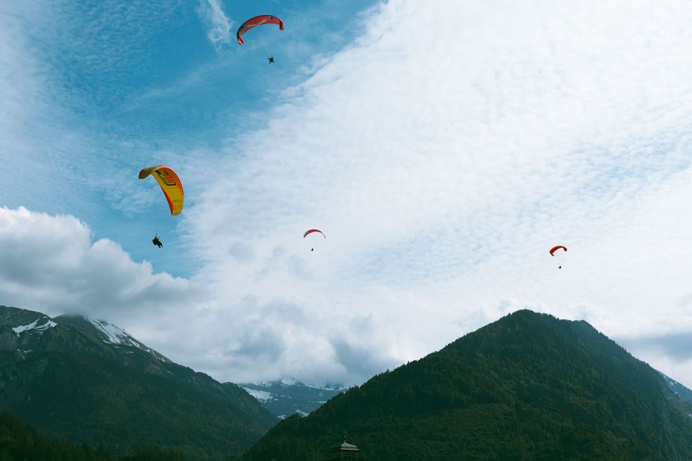 Colorful paragliders soar above snow-capped mountain peaks against a bright blue sky with scattered clouds.