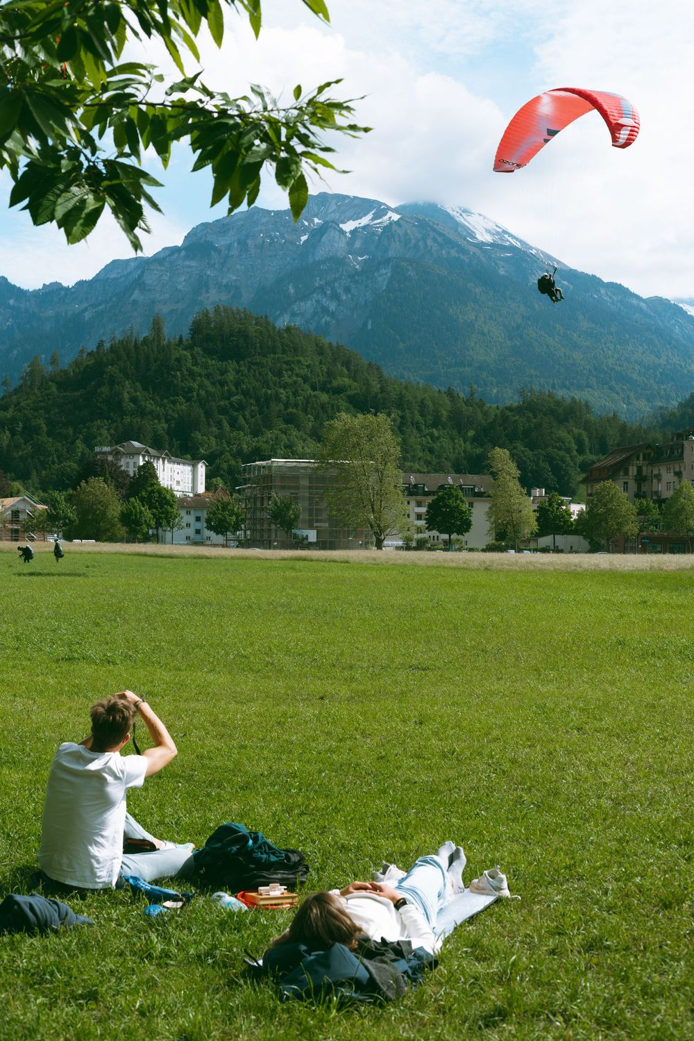 A paraglider soars over a grassy field near mountains while someone watches from below in the Alpine landscape.
