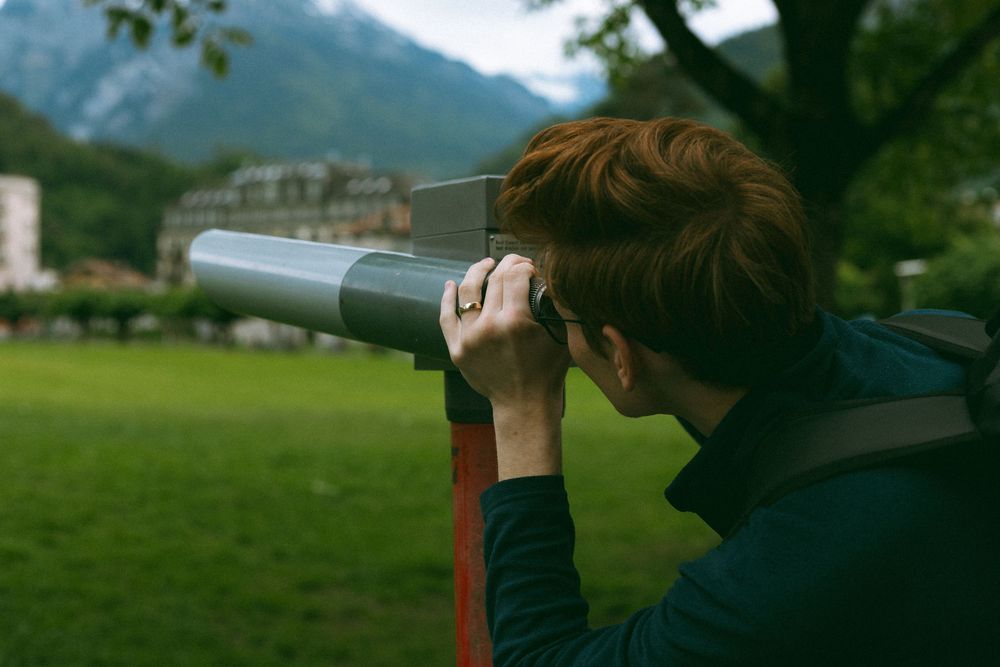 Person looks through a telescope in a mountain valley with green meadow and mountainous terrain in the background.