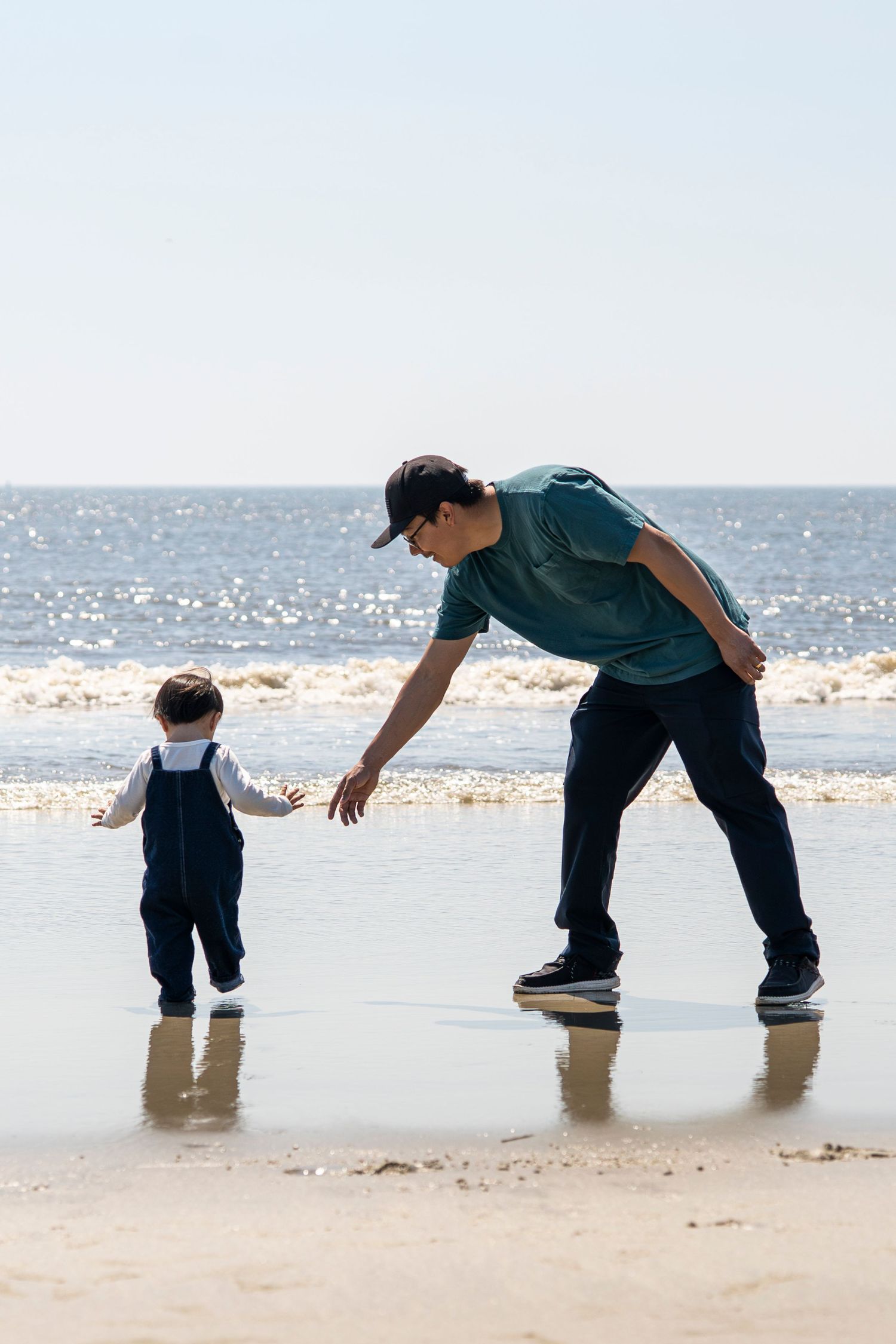 Father bending down to hold toddler's hand on sunny beach shoreline with ocean waves in background.