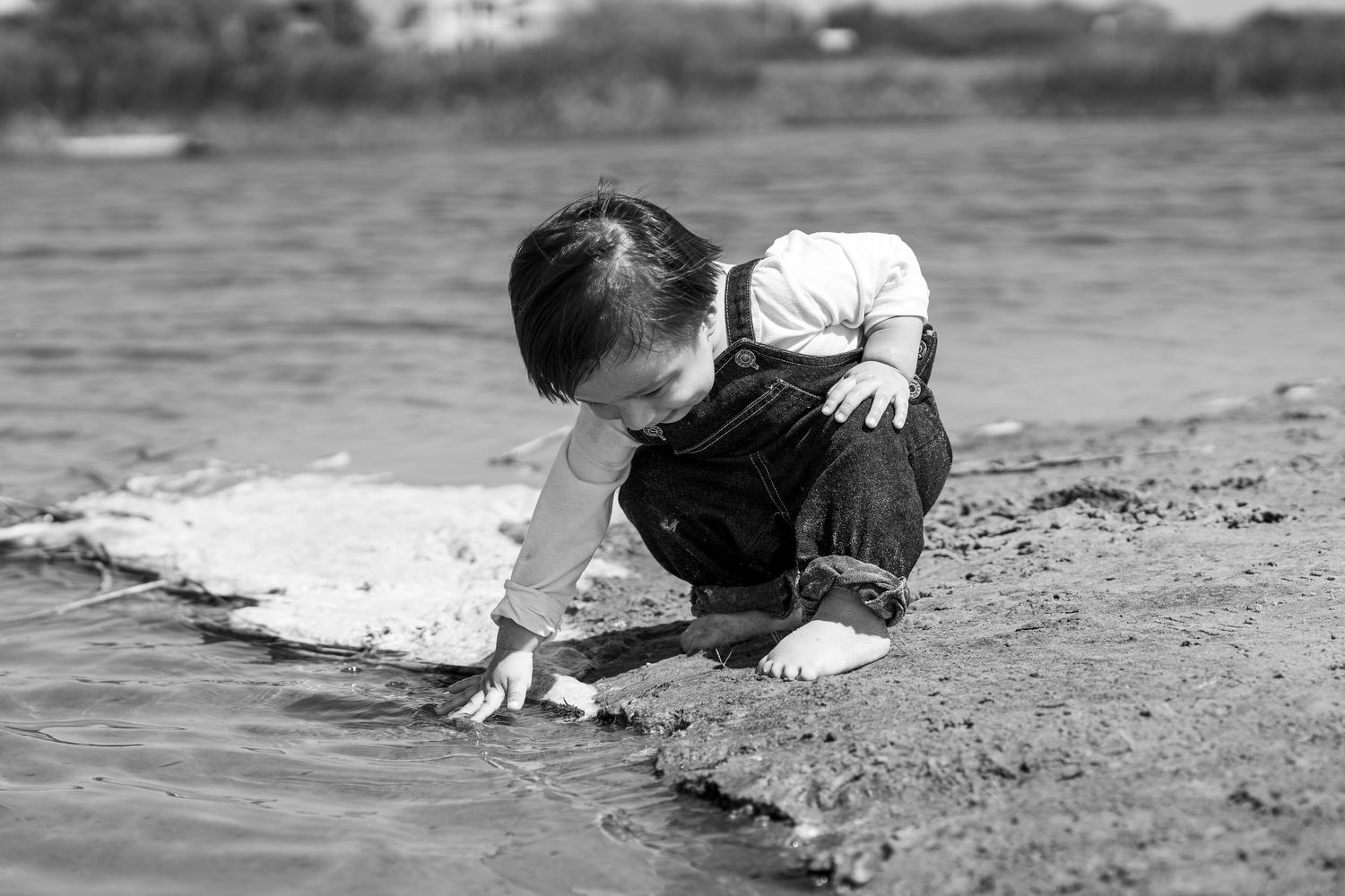 Black and white photo of a toddler playing in shallow water at a sandy beach shoreline.