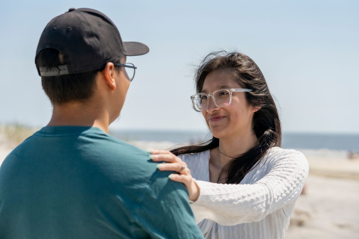 A young woman with glasses smiles at a man on a sunny beach, gently placing her hands on his shoulders.