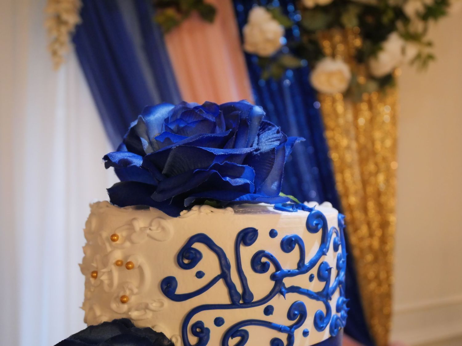 Elegant wedding cake decorated with blue roses and gold accents against blue and gold backdrop.