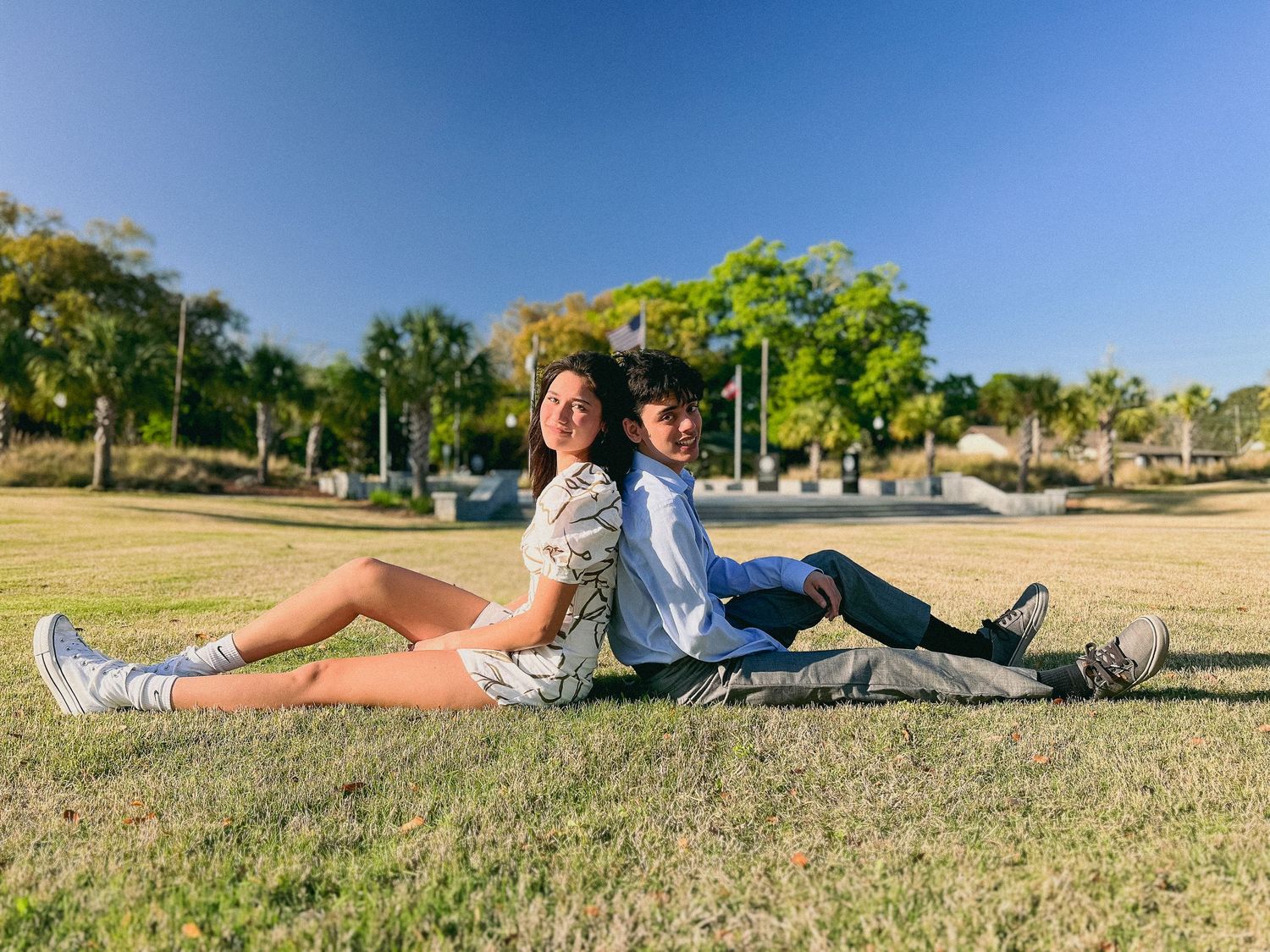 People sitting back to back on grass in a sunny park setting.