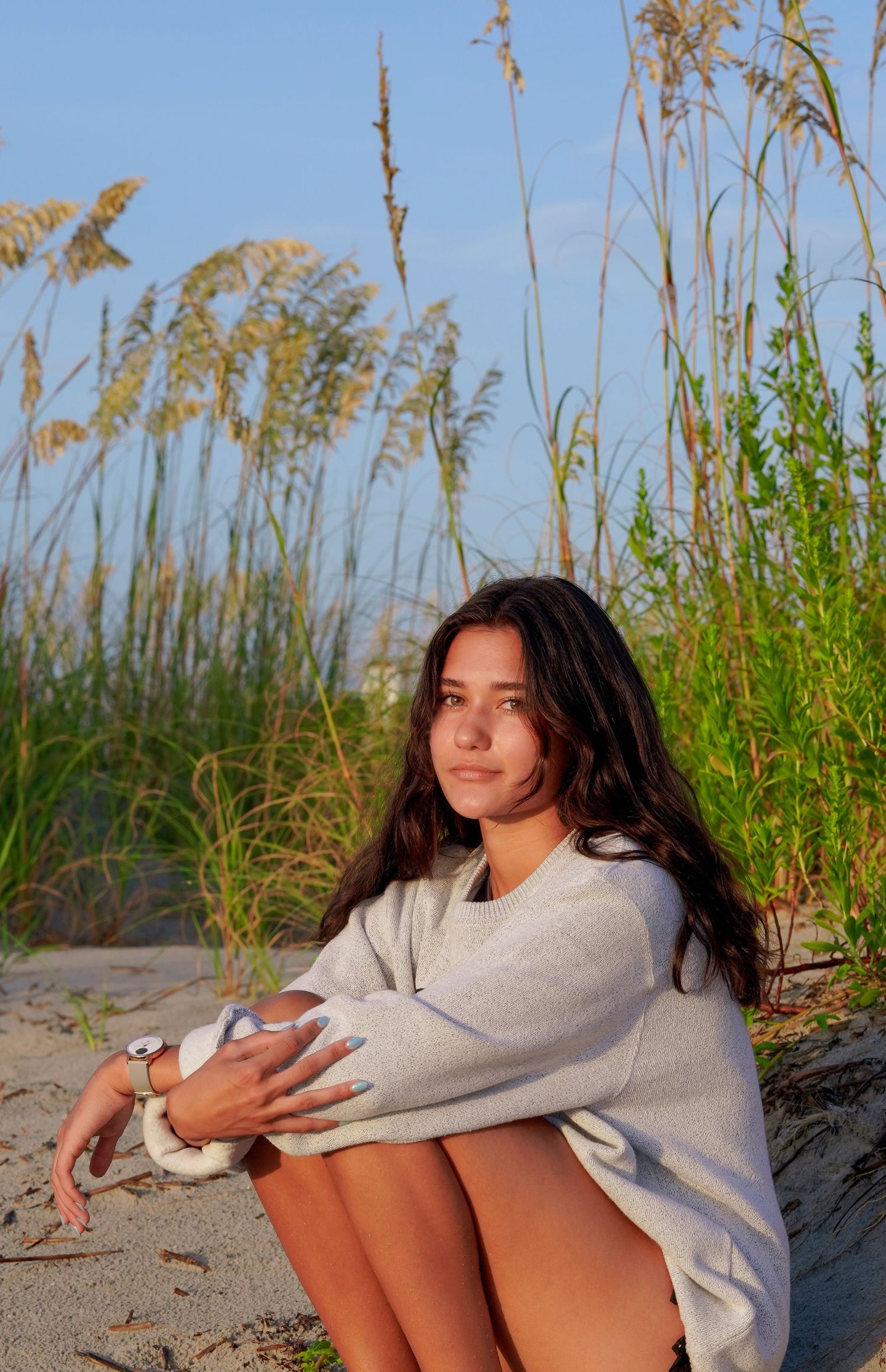 Person relaxing on sandy beach wearing oversized gray sweatshirt amidst tall sea grasses at golden hour.