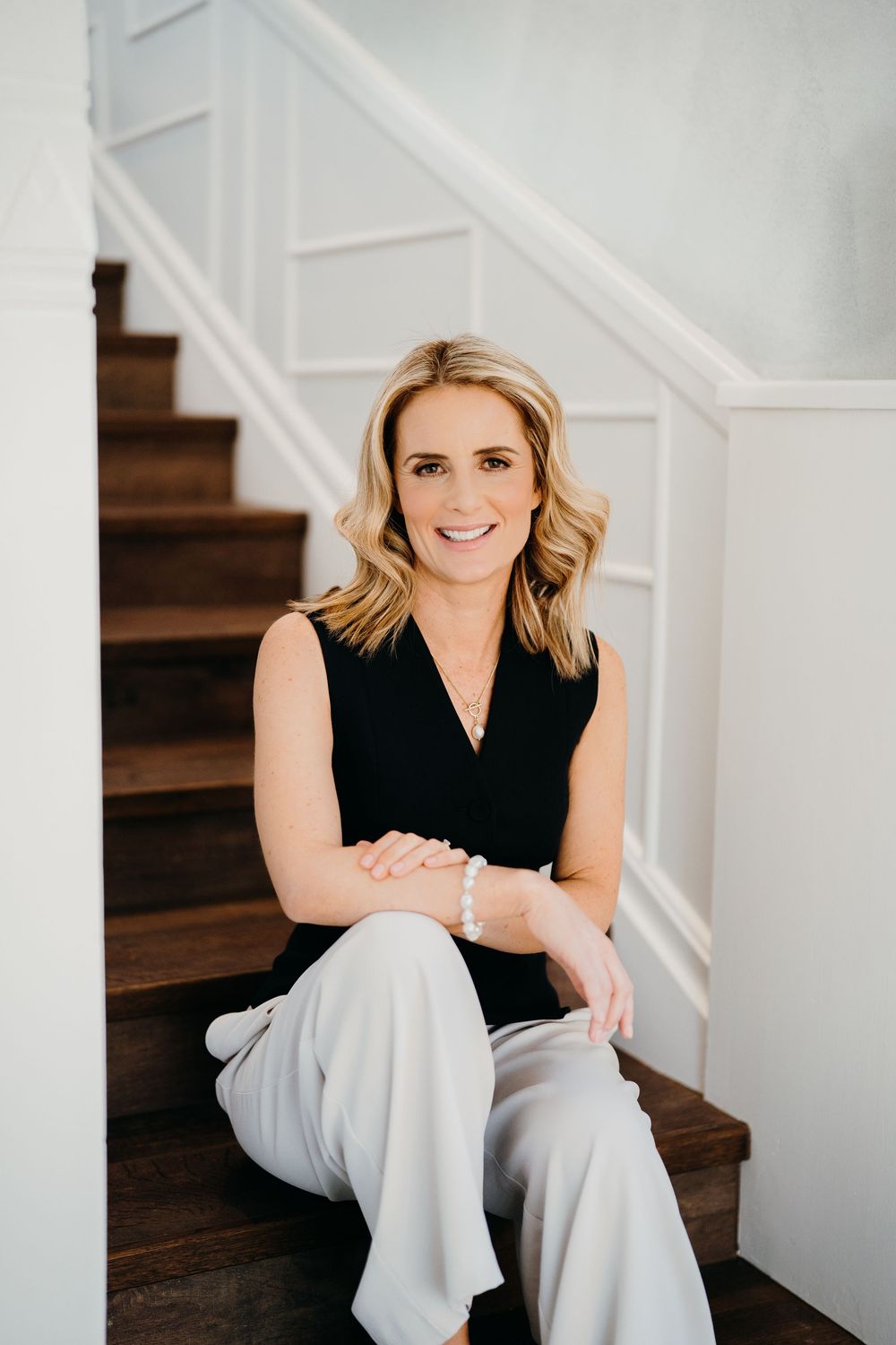 Professional lifestyle portrait of someone sitting on wooden stairs wearing a black sleeveless top and light colored pants.