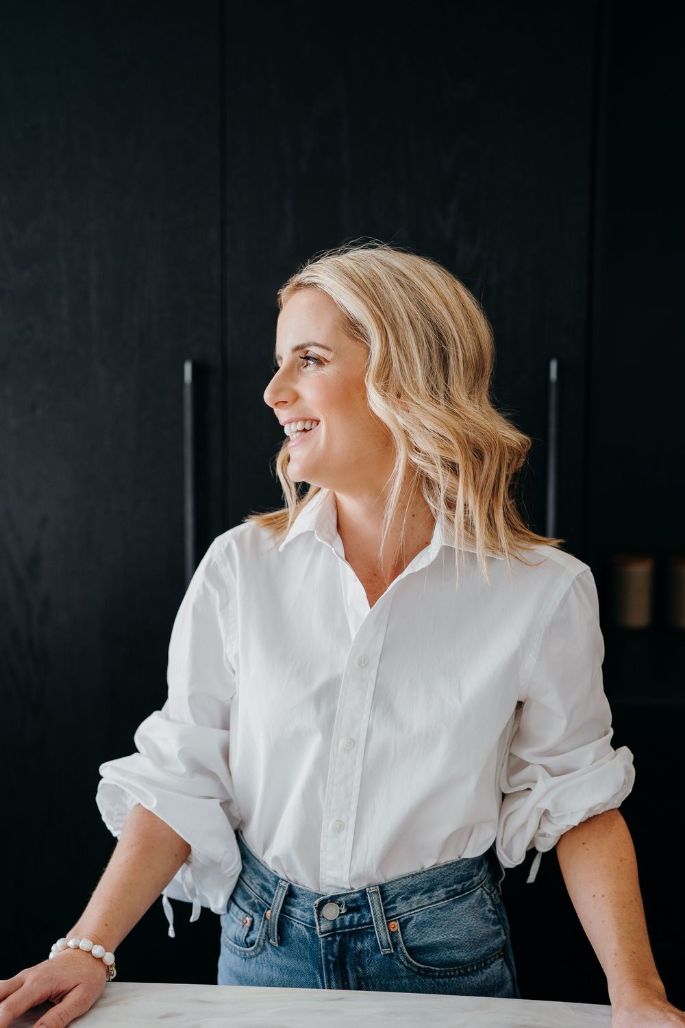 Person in white button-down shirt and jeans laughs while looking to the side against dark background.