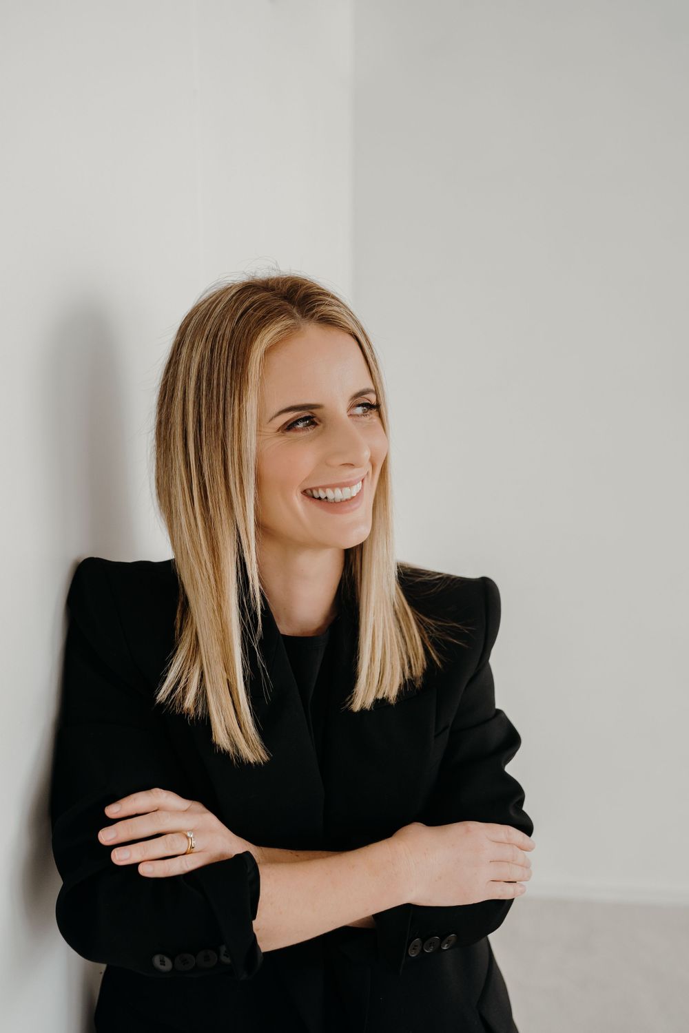 Professional portrait of someone in a black blazer standing against a white wall with a bright smile.