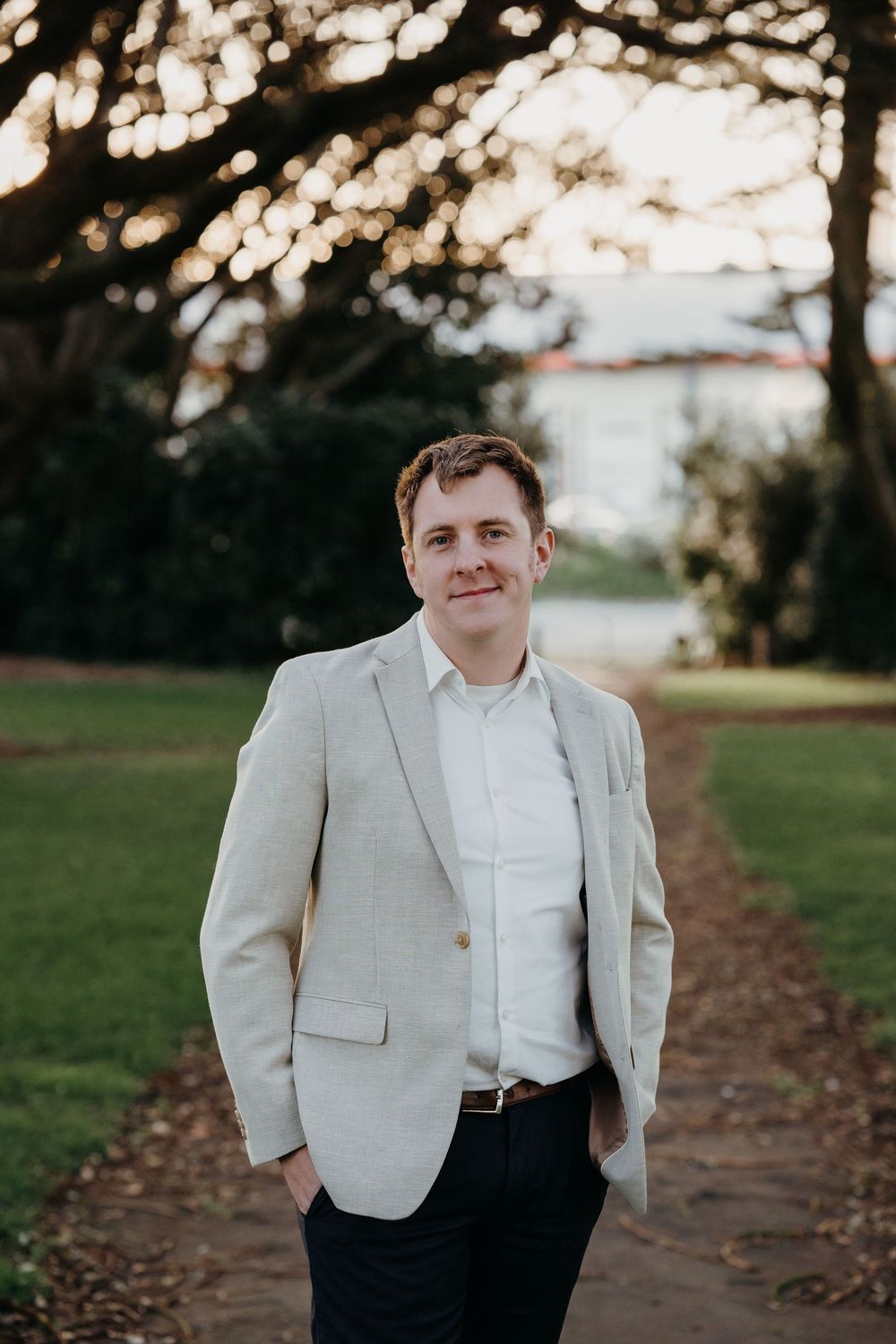 Professional portrait of someone in a light gray blazer and white shirt standing on a tree-lined path at golden hour.