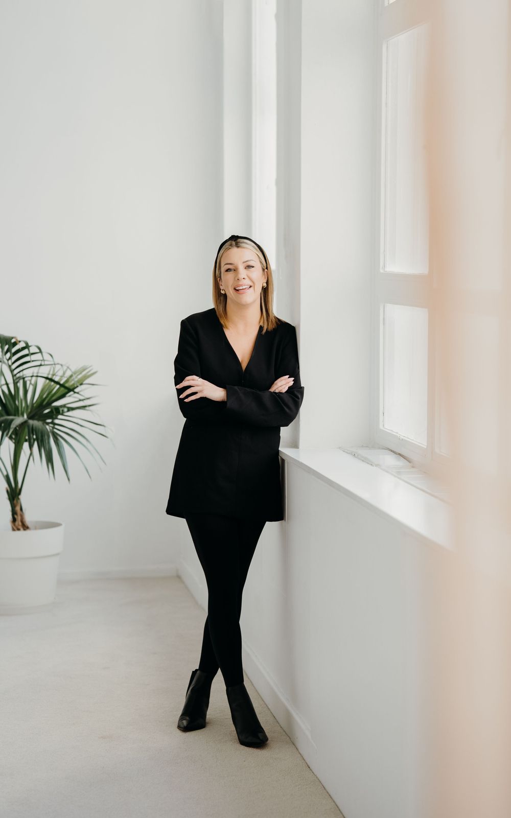 Professional in sleek black business attire poses confidently against white wall with minimalist decor.