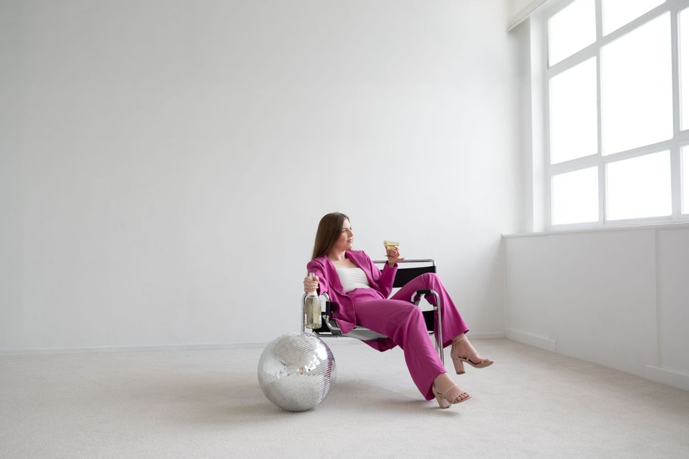 Person in bright fuchsia satin pajamas sits on white floor with silver disco ball in minimalist room near window.