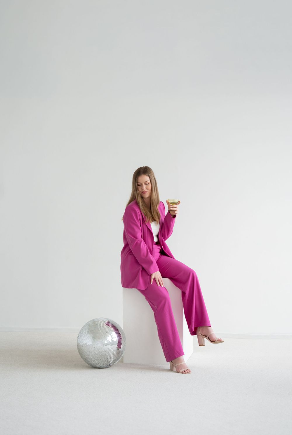 Person in bright pink pantsuit sitting casually next to large disco ball on white background holding champagne glass.