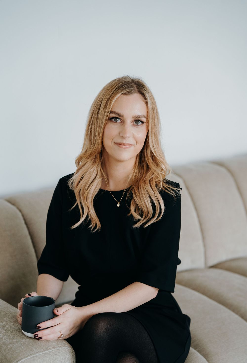 A person wearing a black dress sits on a cream colored couch holding a dark coffee mug.