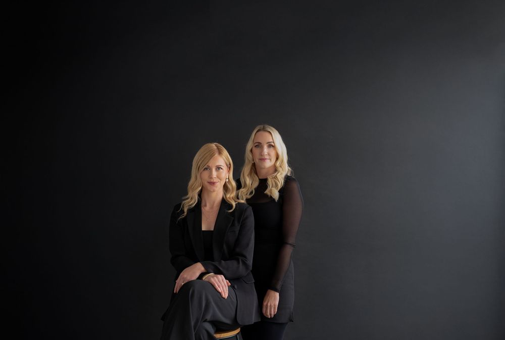 Two women in black outfits pose against a dark background in a professional studio portrait.
