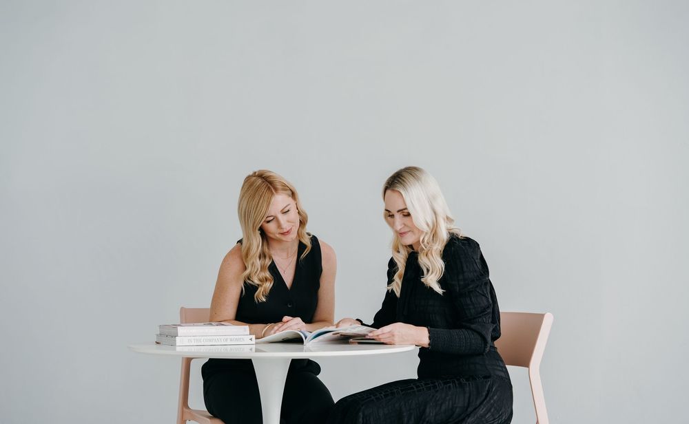 Two business colleagues in black attire sit at a white table reviewing documents together against a light gray background.