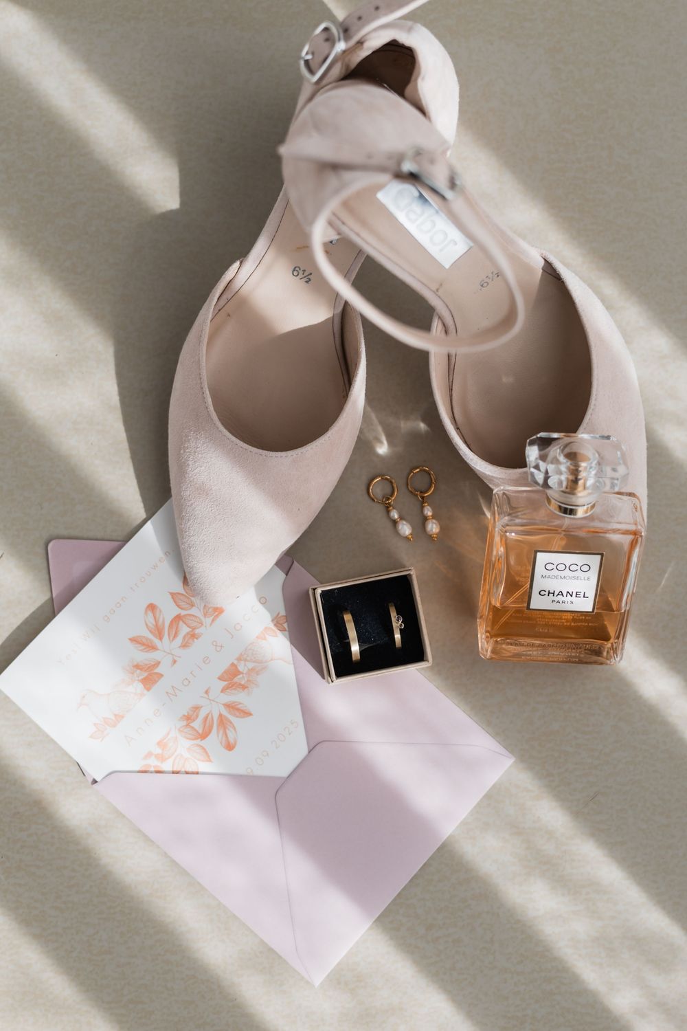 Wedding accessories including beige heels, perfume bottle, earrings and invitation card arranged on white surface in natural light.