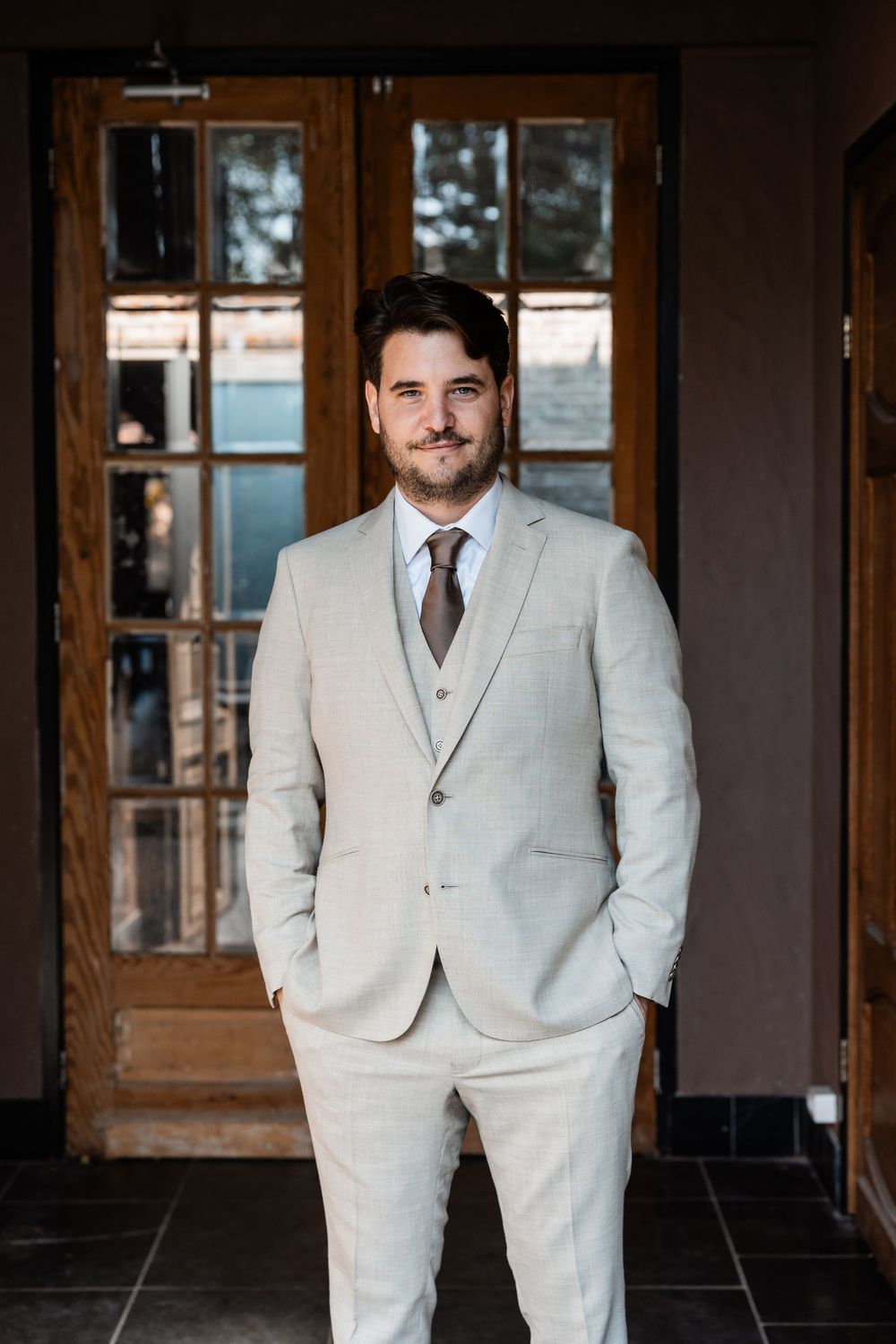A groom in a light gray suit and vest poses in front of wooden doors at a wedding venue.