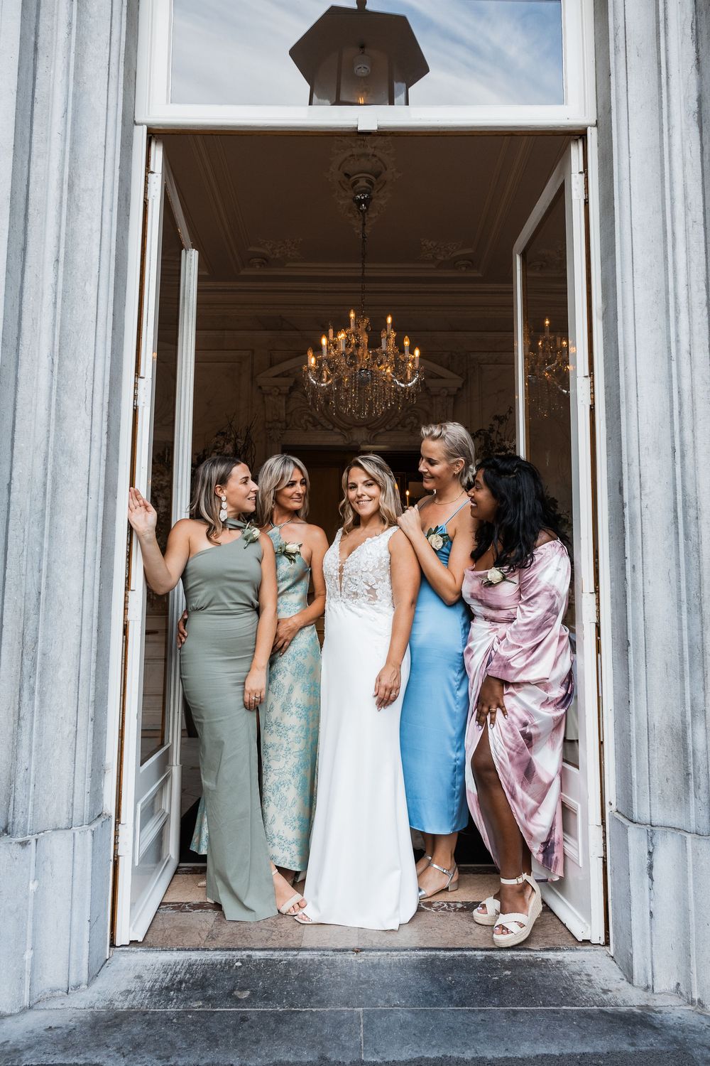 A group of women in formal dresses laugh together while sitting in a row along white columns at an elegant venue.