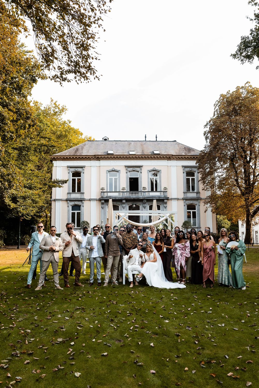 Wedding party gathers for group photos on a lawn in front of an elegant white mansion on an autumn afternoon.