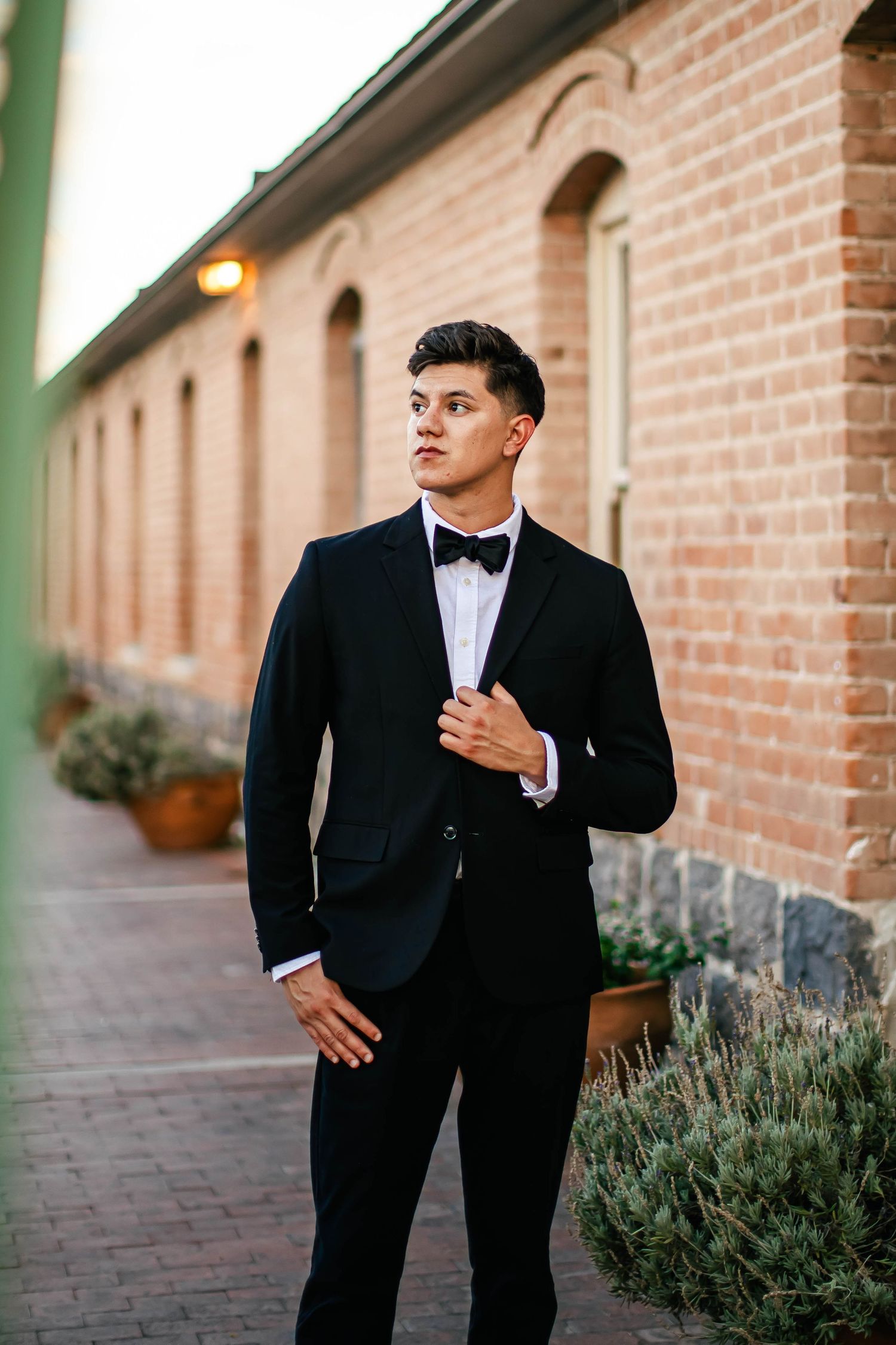 Groom in black tuxedo poses against brick wall with greenery.
