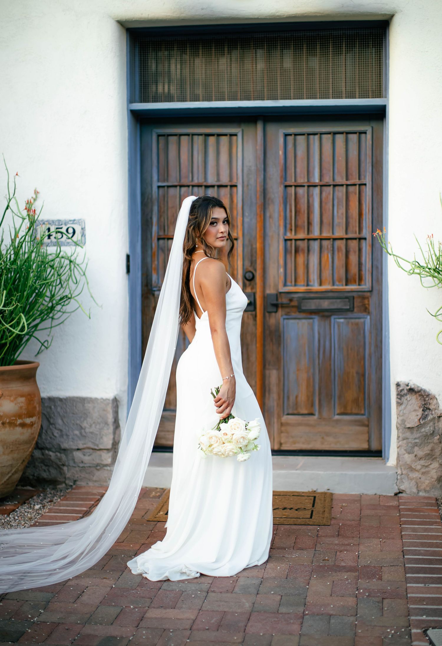 Bride in flowing white dress with long veil poses by rustic wooden doors.
