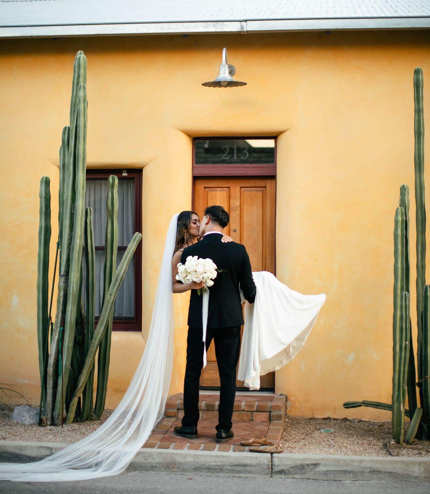 Wedding couple poses against yellow adobe wall with tall desert cacti creating geometric shadows.