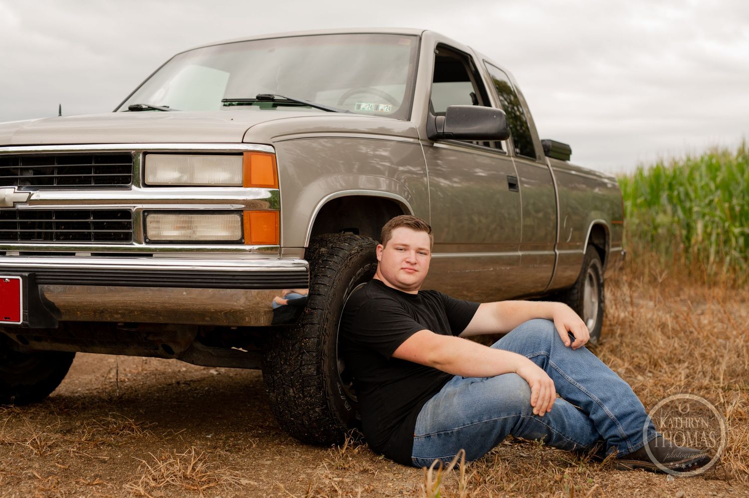 Someone poses casually while sitting against the wheel of a silver Chevy pickup truck near a cornfield.