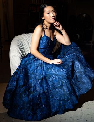 A woman in an elegant royal blue lace evening gown poses gracefully against a dark background.