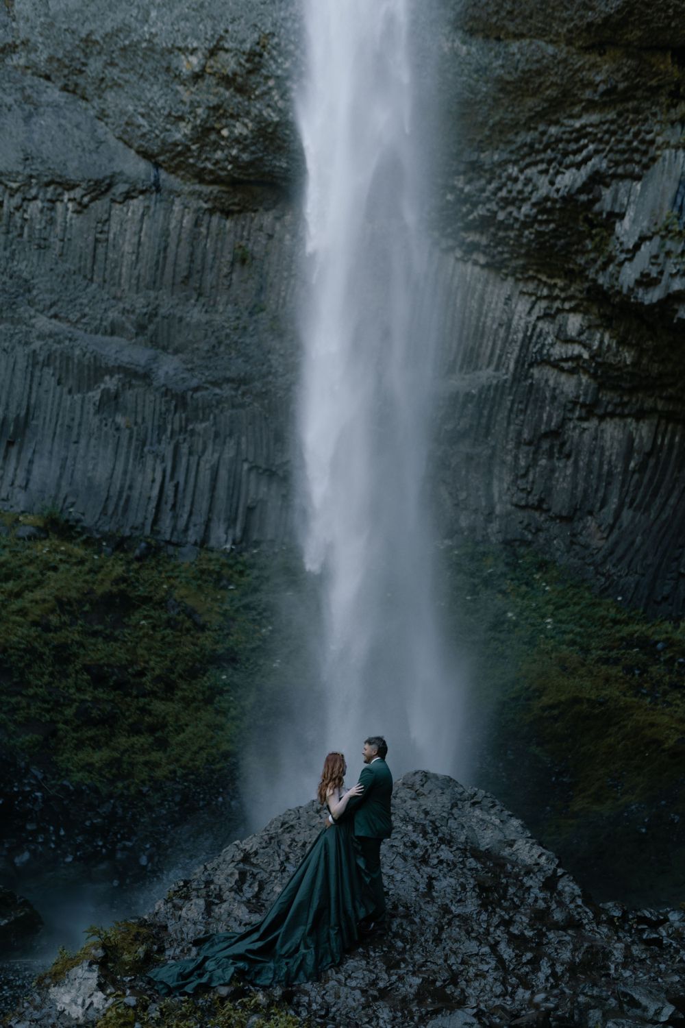 Majestic waterfall cascades down dark basalt cliffs as figures in dark clothing pose on rocky outcrop.
