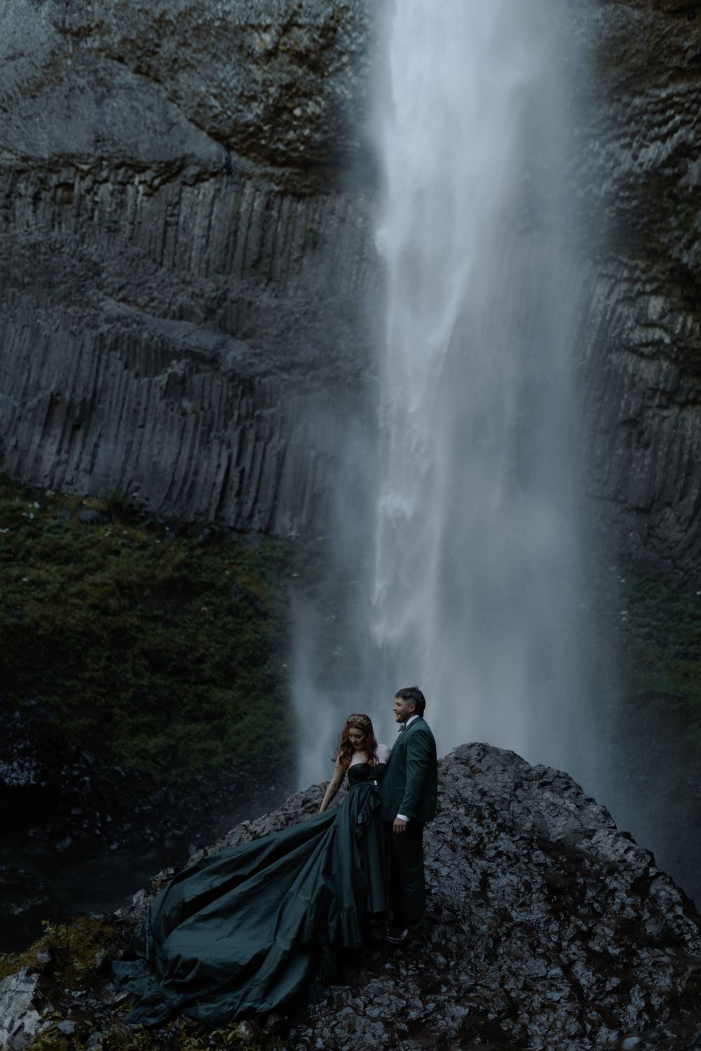 Two silhouettes stand against a powerful waterfall backdrop on a rocky ledge.