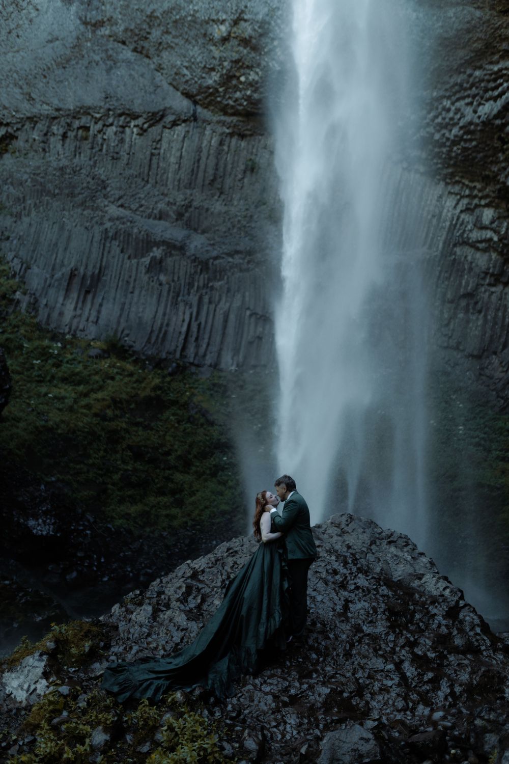 Couple in dark attire embraces near base of towering waterfall.