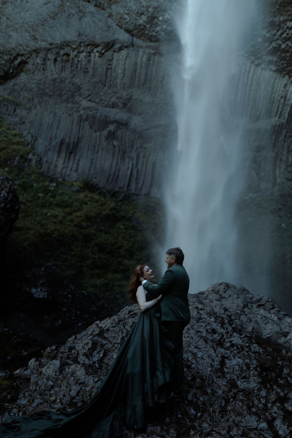 Two people in dark clothing pose dramatically by massive waterfall.