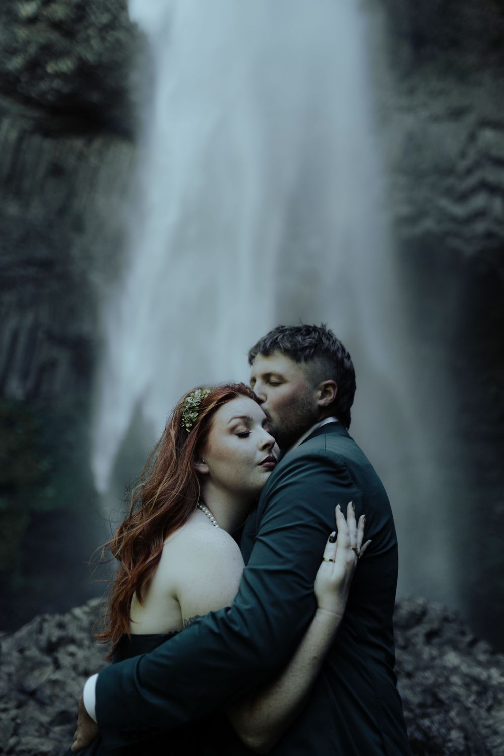 Intimate moment captured against misty waterfall background in dark tones.